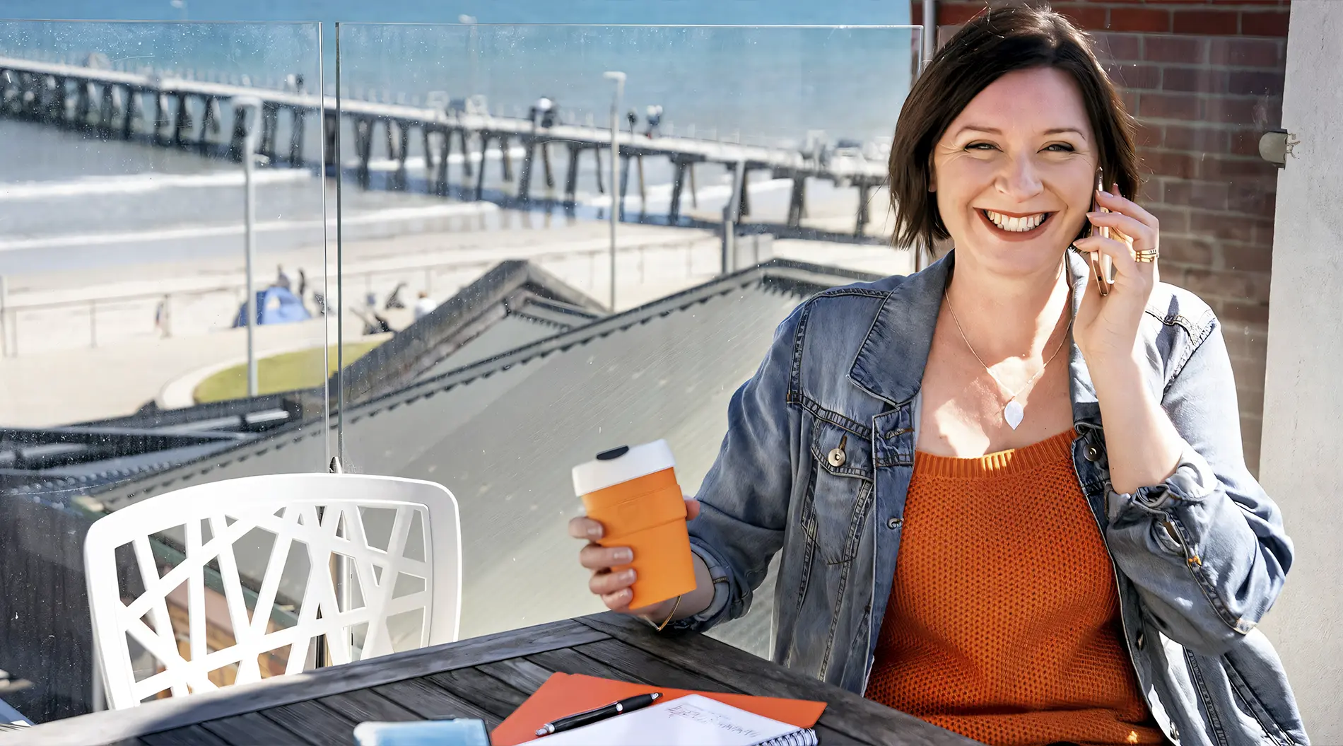 Tammy Edwards smiling on the phone with a coffee, on a balcony overlooking the beach and jetty